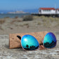 Sunglasses with blue-green lenses and matte-black arms. Handstitched sunglasses sleeve behind sunglasses. Golden Gate Beach in the background.