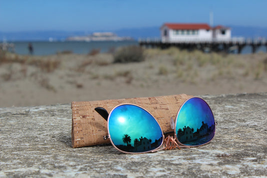 Sunglasses with blue-green lenses and matte-black arms. Handstitched sunglasses sleeve behind sunglasses. Golden Gate Beach in the background.