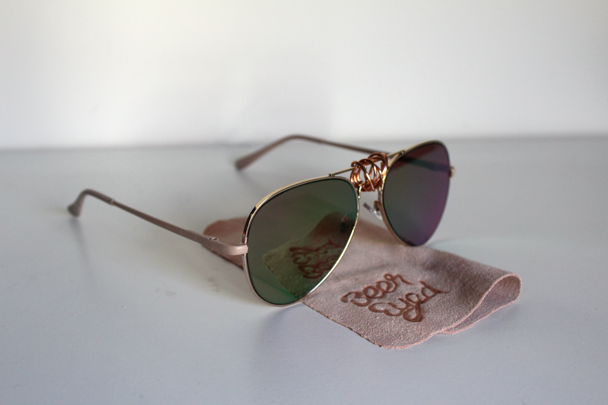 Sunglasses with rose-gold lenses and matte-rose arms.  Beer Eyed sunglasses cloth under sunglasses. Left-view