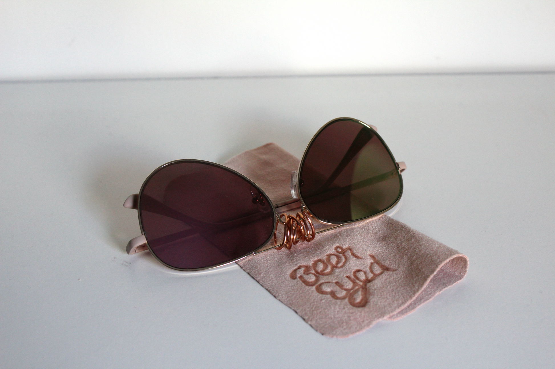 Sunglasses with rose-gold lenses and matte-rose arms.  Beer Eyed sunglasses cloth under sunglasses. 